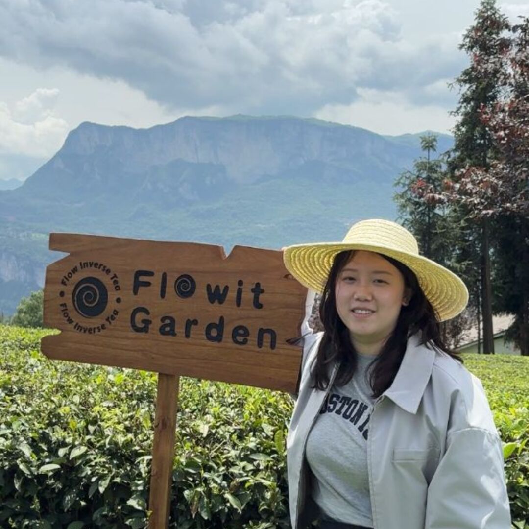 Tea farmer in a sun hat standing next to organic tea garden sign with mountains in the background.